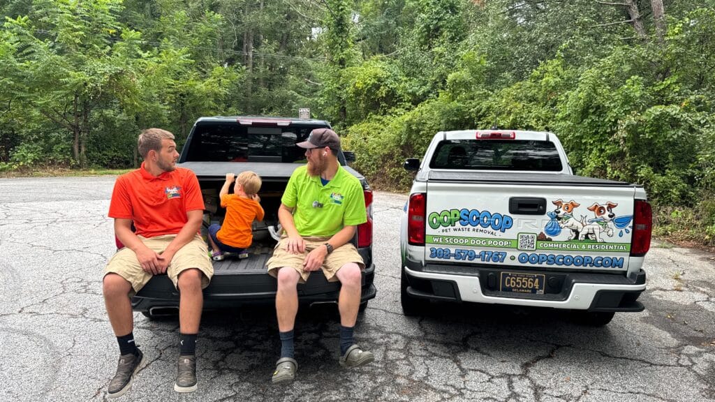 Oop Scoop technicians and a child sitting on a truck tailgate next to an Oop Scoop branded service truck for pet waste removal in Delaware and Maryland.